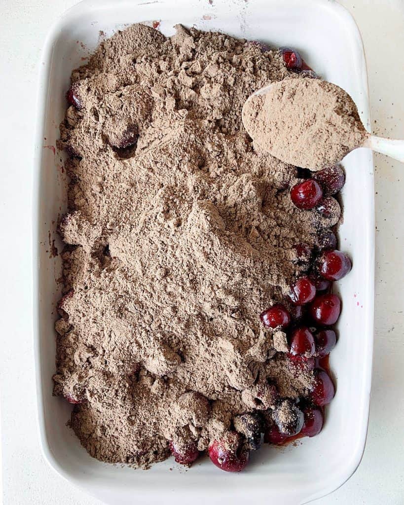 Sprinkling chocolate cake mix with a spoon over cherry filling in a baking dish.
