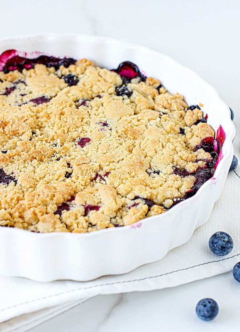 Partial view of round white dish with bluberry crumb cake, white surface, loose blueberries
