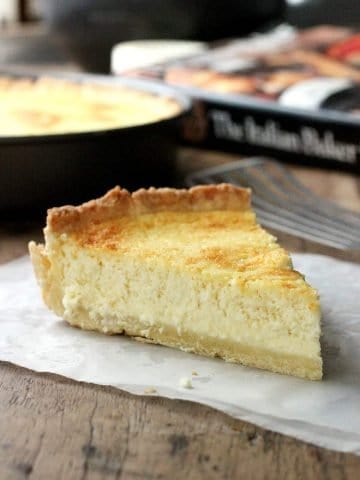 Single slice of ricotta pie on parchment over wooden table, with book, pan in background