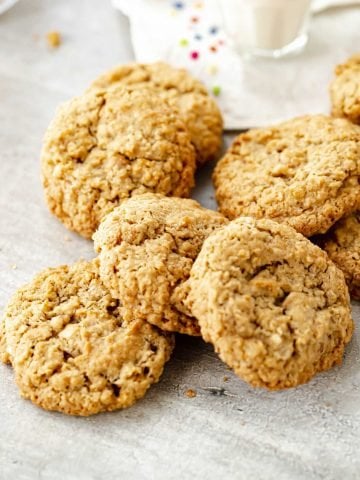 Several oatmeal cookies heaped together on grey surface, color dotted white linen in background.
