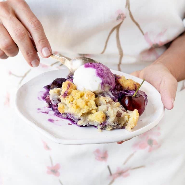 Hands holding a white plate with cherry cobbler serving. White and cherry blossoms background.